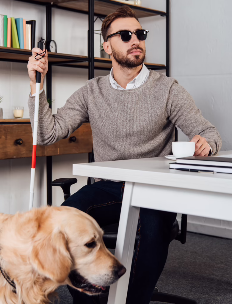 A man with visual impairment sits at a desk, wearing sunglasses and holding a white cane and a coffee cup, with a Golden Retriever service dog in the foreground.